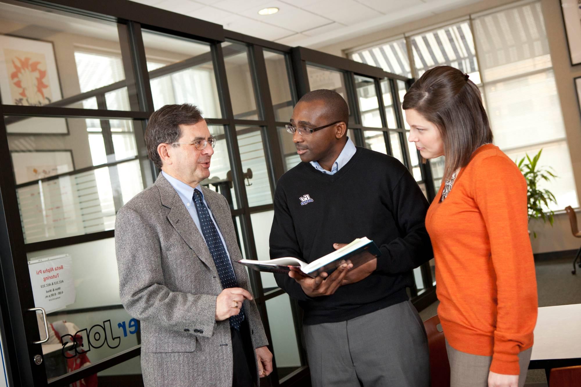 Three faculty members engaged in a discussion while standing and referencing a book